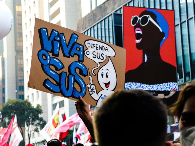 Imagem de uma manifestação na avenida Paulista com foco em um cartaz escrito Viva o SUS com o Zé Gotinha dizendo Proteja o SUS